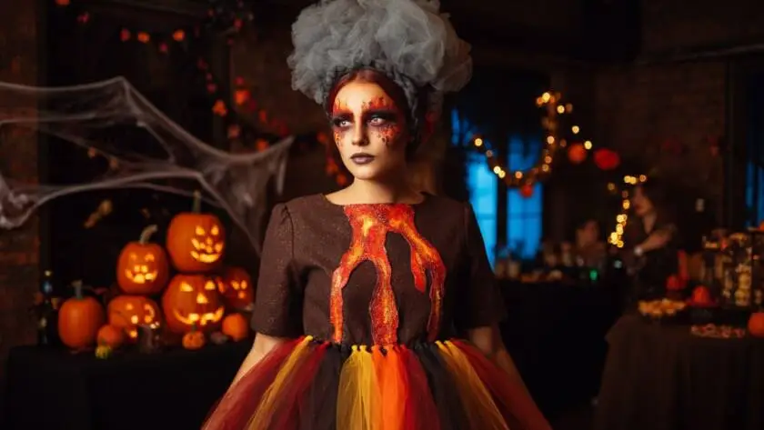 A woman dressed as "Volcano Vickie" at a Halloween party. She wears a dark brown dress resembling a volcano, with a large black fabric volcano structure at her waist. Bright red, orange, and yellow tulle flows down from the volcano, mimicking lava.