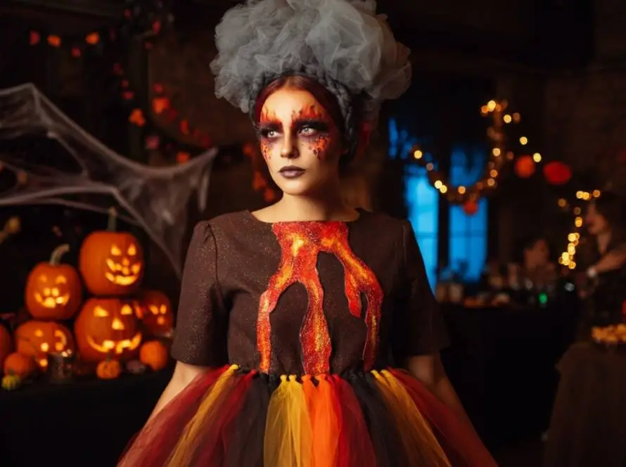 A woman dressed as "Volcano Vickie" at a Halloween party. She wears a dark brown dress resembling a volcano, with a large black fabric volcano structure at her waist. Bright red, orange, and yellow tulle flows down from the volcano, mimicking lava.